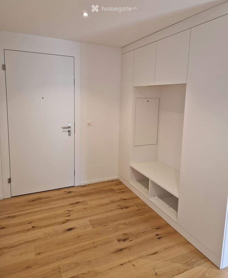 Empty room with light wooden flooring, white walls, built-in white shelving unit, and a closed white door.