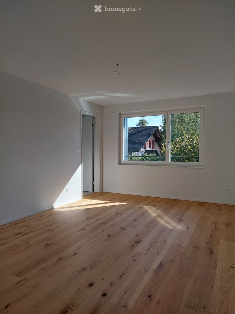 Empty room with light wooden flooring, white walls, and a large window letting in natural light. The room has a door on the left side and visible electrical outlets on the walls.