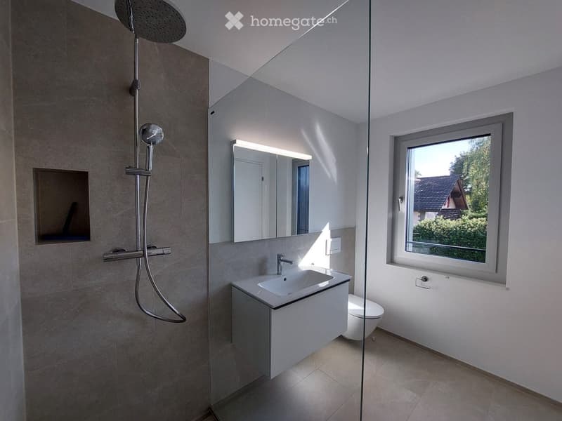 Modern bathroom with a glass-enclosed shower, a white sink with a cabinet, a toilet, and a large mirror. The room has a large window offering a view of the outside.