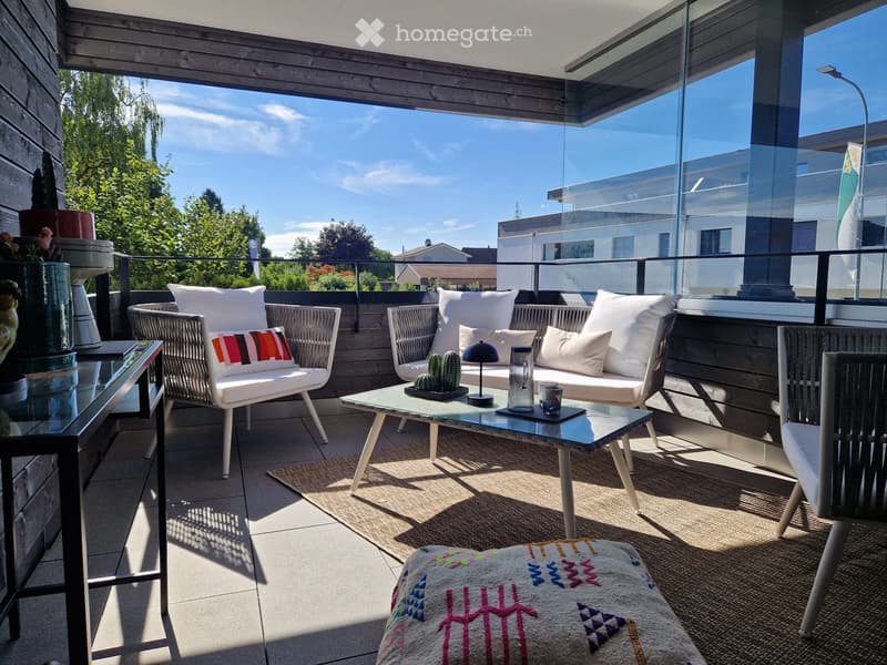 A modern balcony with a sofa, coffee table, chairs, and decorative cushions, featuring a glass railing and a view of greenery.