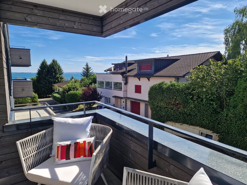 Balcony with wooden decking, white wicker chair, small table, and colorful cushion, overlooking a scenic view of the lake and surrounding houses.
