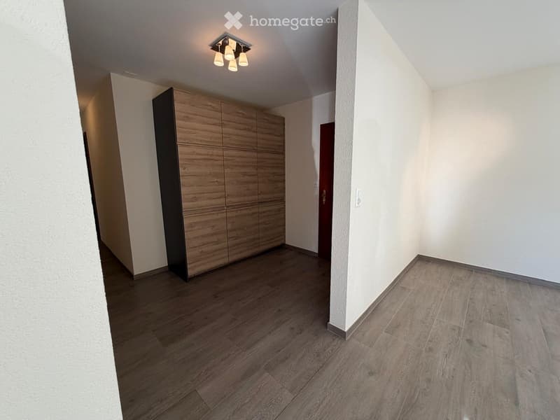 Empty room with wooden flooring, white walls, and a built-in wooden wardrobe. The room has a modern design with recessed lighting and a visible door leading to another area.