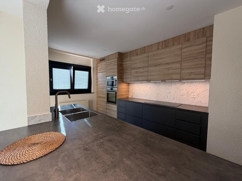 Modern kitchen with wooden cabinets, stainless steel sink, and built-in oven. Features a large window with blinds and a woven mat on the floor.
