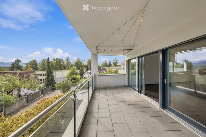 Modern balcony with tiled floor, glass railing, and large sliding glass doors leading to the interior. Offers a scenic view of trees and distant mountains.