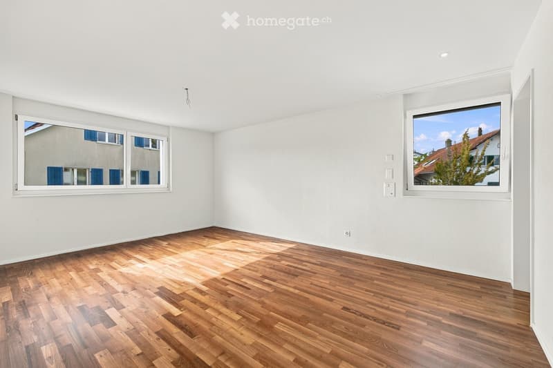 Empty room with wooden flooring and white walls, featuring large windows with views of neighboring buildings and greenery.