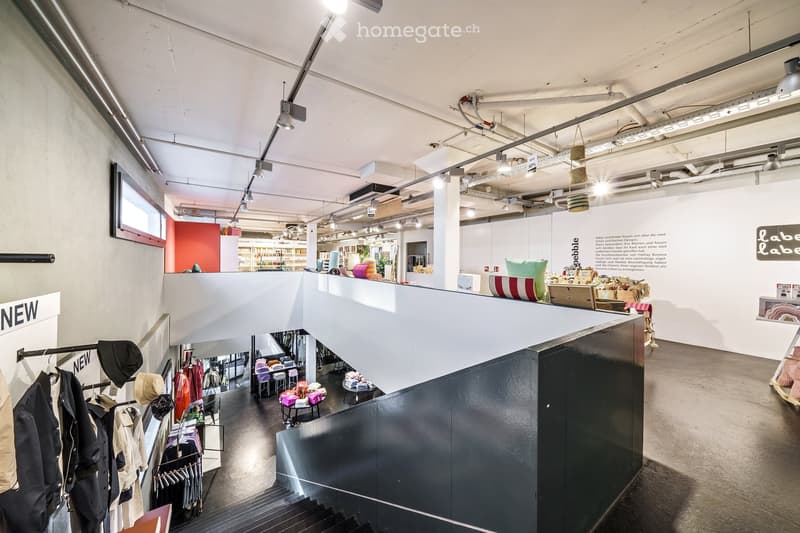 Modern interior space with exposed ceiling beams, hanging lights, and a multi-level layout. Features clothing racks, display shelves, and a staircase leading to an upper level.