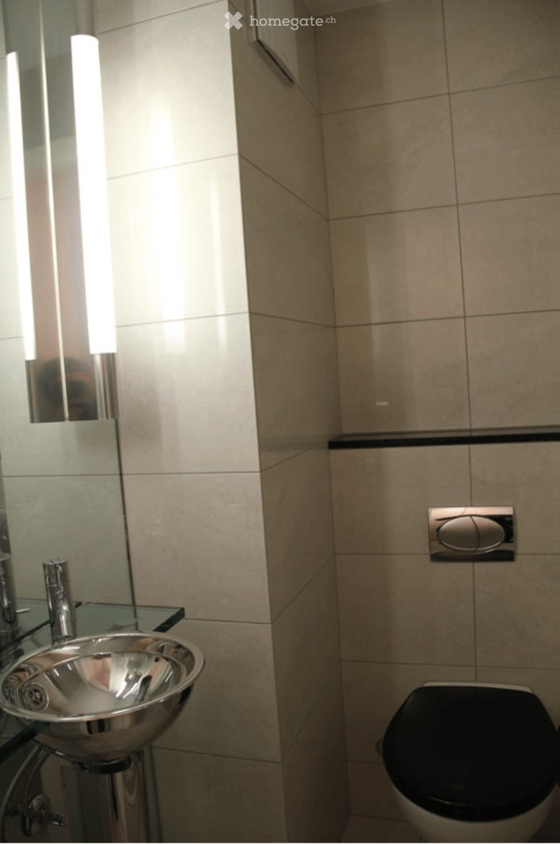 Modern bathroom with beige tiled walls, a sleek metallic sink, a chrome faucet, a large mirror, and a toilet with a black seat.