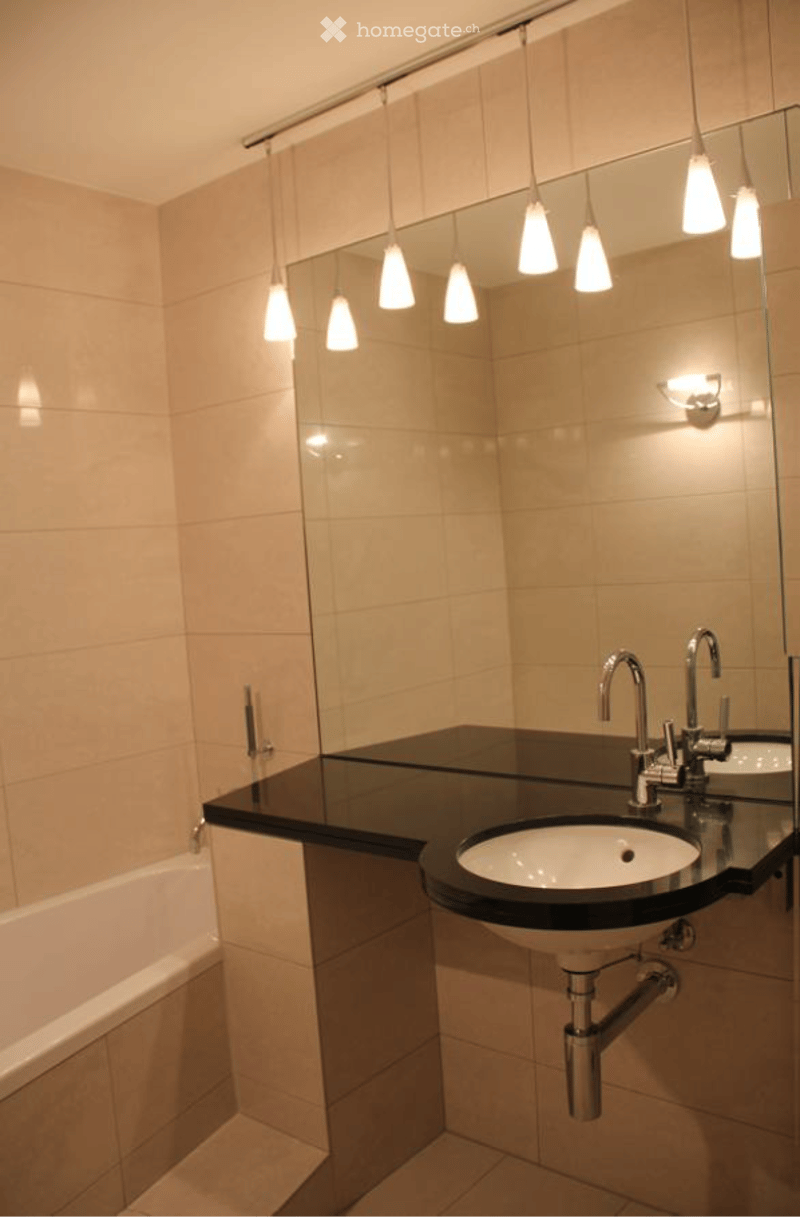 Modern bathroom with beige tiled walls, a black countertop, a white sink, a bathtub, and a large mirror with pendant lights above.