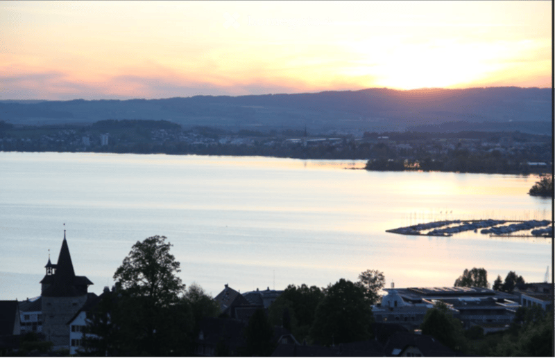 Panoramic view of a lake at sunset, with a town and distant mountains visible in the background.