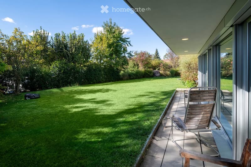 A well-maintained garden with a neatly trimmed lawn, surrounded by trees and shrubs. A wooden deck features lounge chairs and a small table, with a modern house visible in the background.
