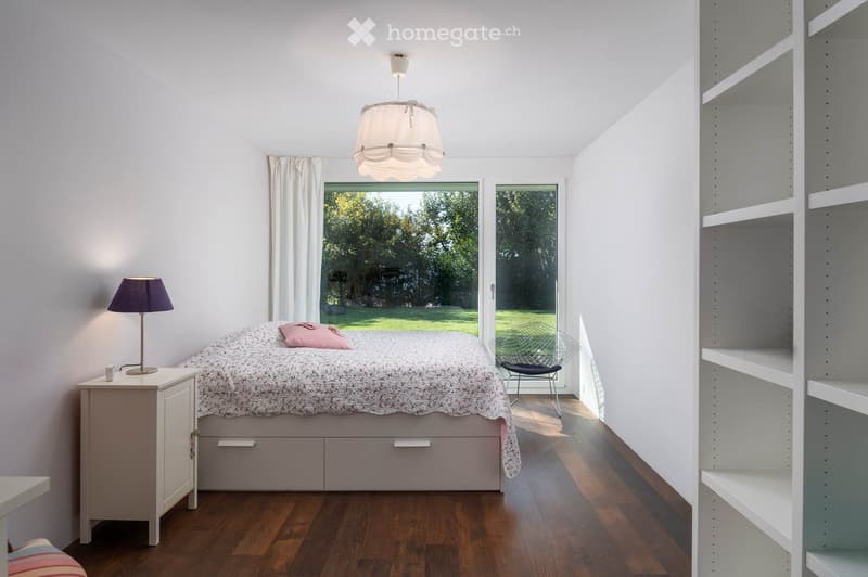 A modern, minimalist bedroom with a wooden floor, white walls, and large windows overlooking a green lawn. The room features a bed with a patterned duvet, a nightstand with a lamp, a built-in shelf unit, and a sleek chair near the window.