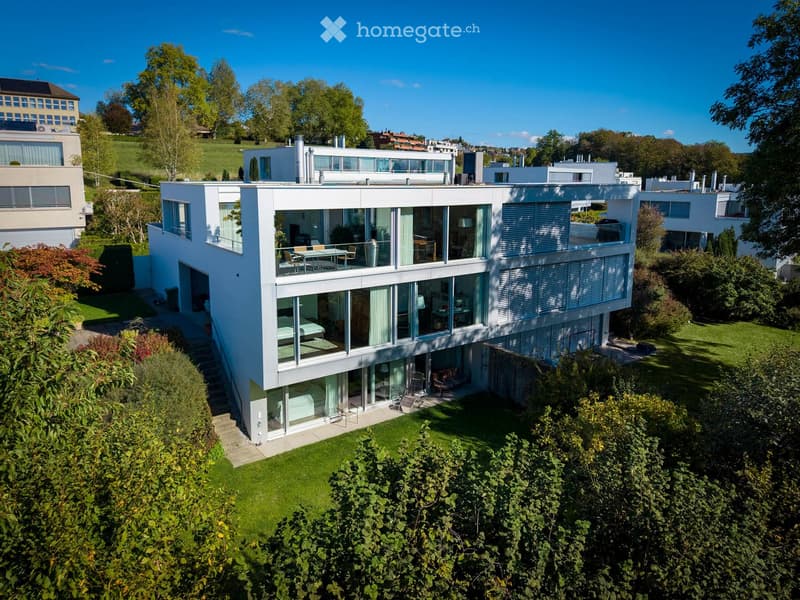 Modern two-story house with large glass windows, a raised ground floor, and a well-maintained garden.