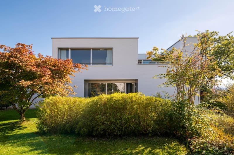 Modern two-story house with white siding, large windows, and a well-maintained garden with shrubs and trees.