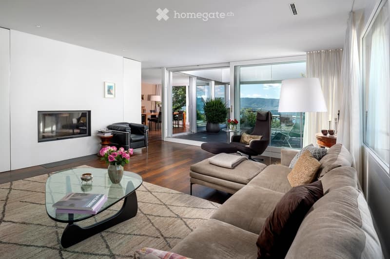 Modern living room with large windows offering a scenic view, featuring a beige sectional sofa, glass coffee table, armchair, ottoman, floor lamp, fireplace, and decorative vase with flowers.