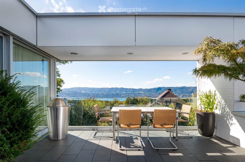 Modern balcony with a glass railing, offering a scenic view of a lake and mountains. It features a small dining set with a table and four chairs, along with potted plants and a trash bin.