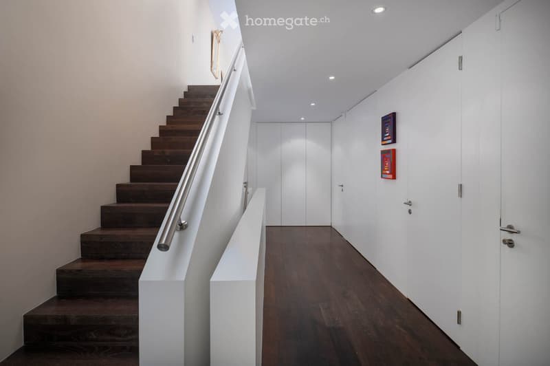 Modern hallway with wooden flooring, white walls, and a staircase with dark wooden steps and a metal railing. Two framed artworks are displayed on the wall.