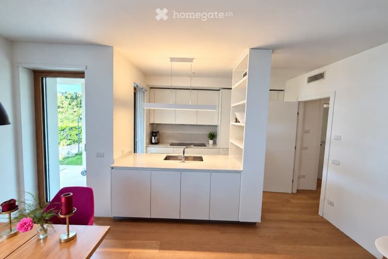 Modern, open-plan kitchen and living area with white cabinetry, a central island, and a small seating area featuring a purple chair and a wooden table with decorative items.