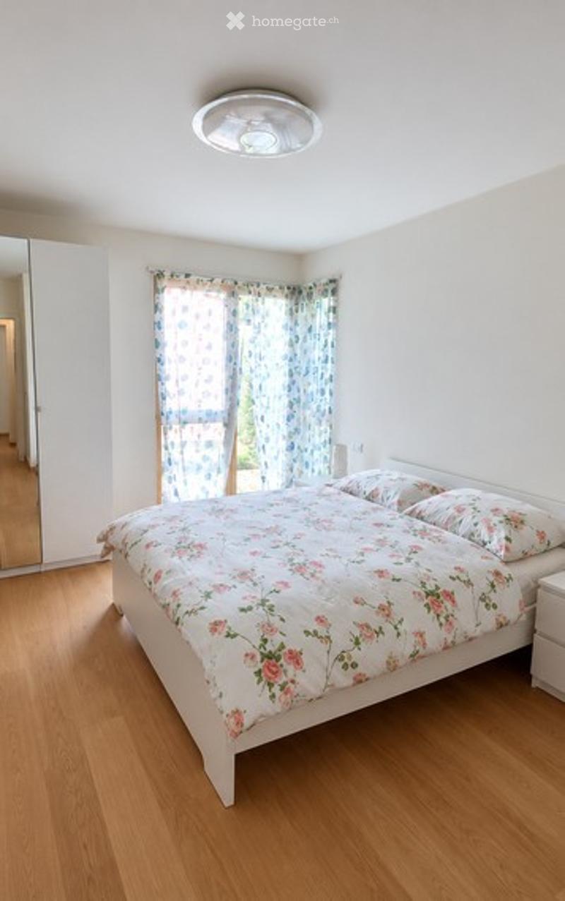 Bright bedroom with wooden flooring, a large bed with floral bedding, a nightstand, and a mirror on the wall. Natural light filters through a window with blue floral curtains.