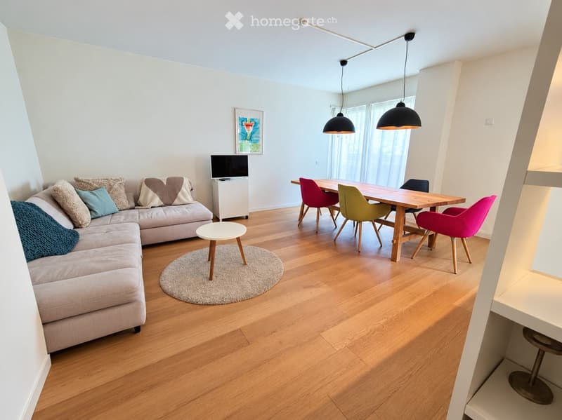 Open-plan living and dining area with light wood flooring, a grey sectional sofa with colorful cushions, a small round coffee table, a dining table with colorful chairs, and a television on a stand.