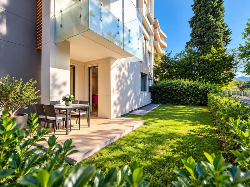 A modern outdoor patio area with a glass-enclosed balcony, a small dining set including a table and chairs, and a flower pot with white flowers. The area is surrounded by a well-maintained lawn and lush greenery.