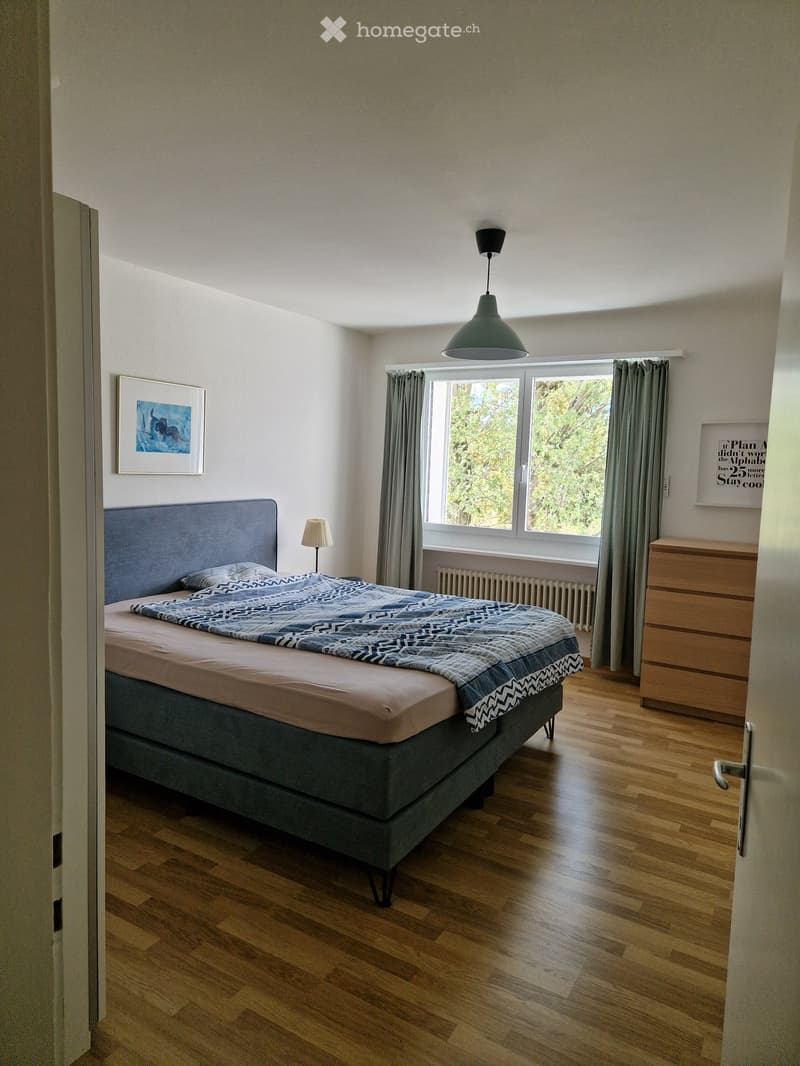 Modern bedroom with wooden flooring, large window with grey curtains, grey upholstered bed with patterned bedding, nightstand with lamp, picture frames on walls, and a chest of drawers.