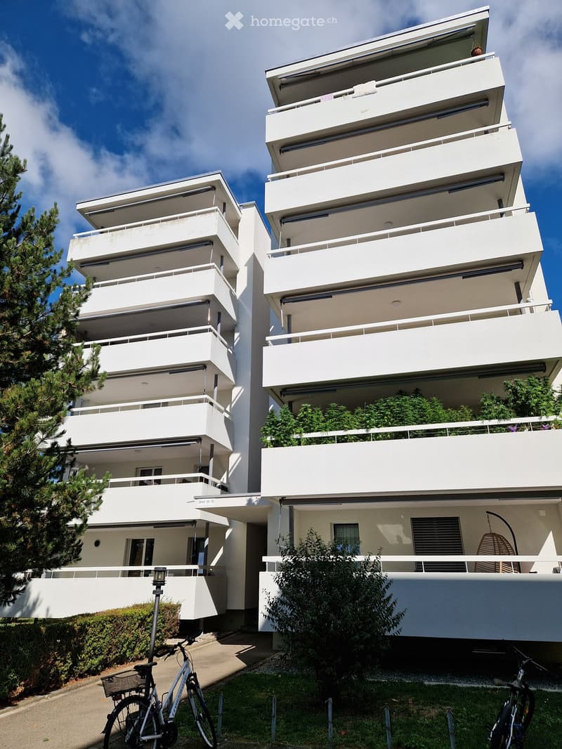 Modern white apartment building with multiple balconies and greenery, bicycles parked in front.