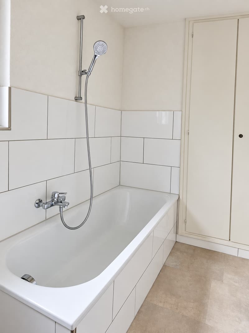 Modern bathroom with a white bathtub, chrome faucet, and handheld showerhead. White tiled walls and light-colored flooring.