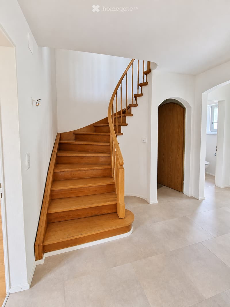 Spacious hallway with a wooden staircase featuring a curved design and a wooden handrail. The floor is tiled, and the walls are painted white. A closed wooden door leads to another room, and a small section of a bathroom is visible in the background.