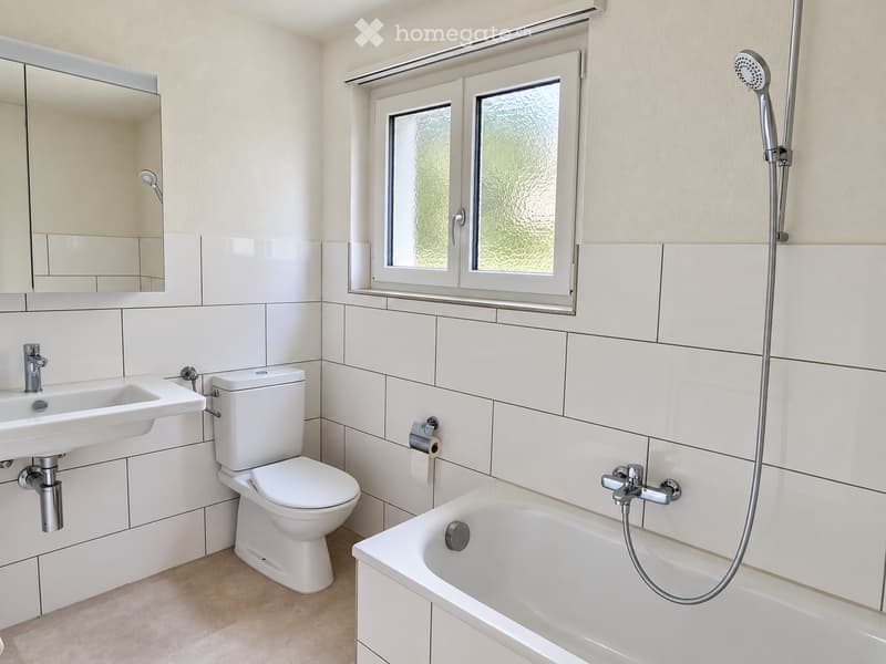 Modern bathroom with white tiles, a sink, toilet, bathtub, and showerhead. Features a large mirror and a window with frosted glass.