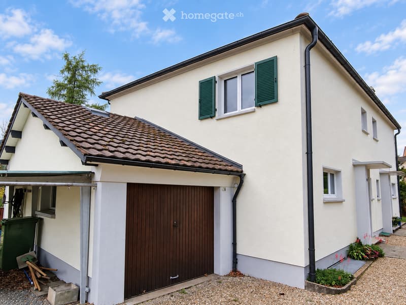 Two-story house with white exterior, green shutters, and a brown garage door. The roof is tiled, and there is a small garden area with plants and flowers near the entrance.