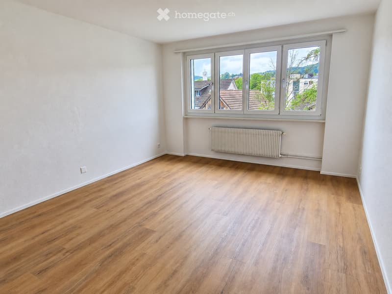 Empty room with wooden flooring, white walls, and large windows offering a view of the neighborhood.
