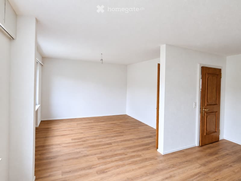 Empty room with light wooden flooring and white walls, featuring a wooden door and a small built-in shelf on the left side.
