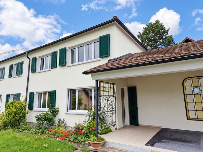 Two-story house with white walls, green shutters, tiled roof, and a small garden with colorful flowers and a potted plant near the entrance.