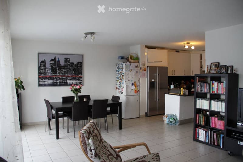 A modern, open-plan living and dining area with a dining table and chairs, a refrigerator, a bookshelf, and a comfortable armchair. The space features white walls, tiled flooring, and a large cityscape picture on the wall.