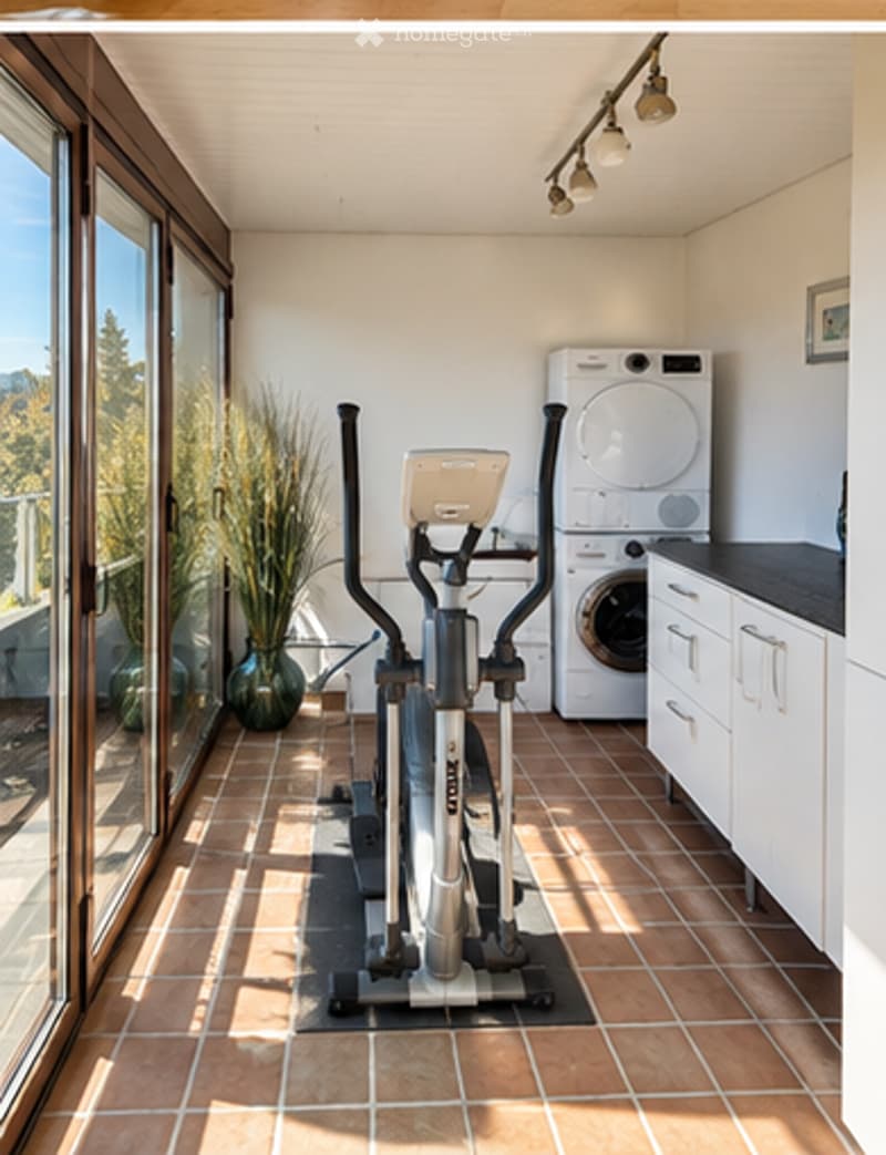 Well-lit room with tiled floor, exercise bike, washing machine, dryer, and decorative vase near large windows offering a scenic view.