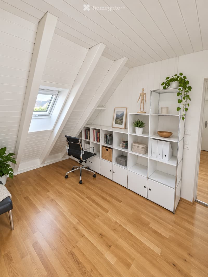 Bright attic room with wooden flooring, slanted white paneled ceiling, large skylight window, and built-in white shelving units holding books, plants, and decorative items.