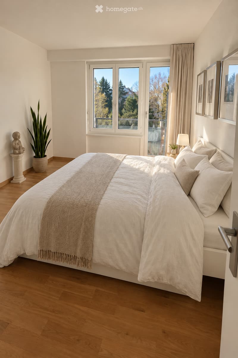 Bright bedroom with wooden flooring, large window with view, bed with pillows and blanket, table lamp, plant, statue, and picture frames on wall.