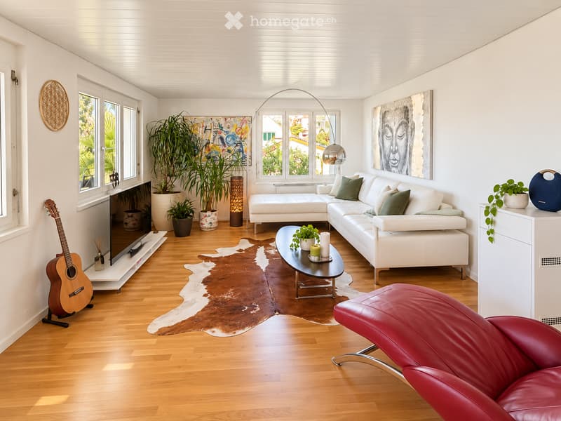 Modern living room with white walls, wooden flooring, large windows, white sectional sofa, coffee table, red leather chair, guitar, screen, and decorative plants.