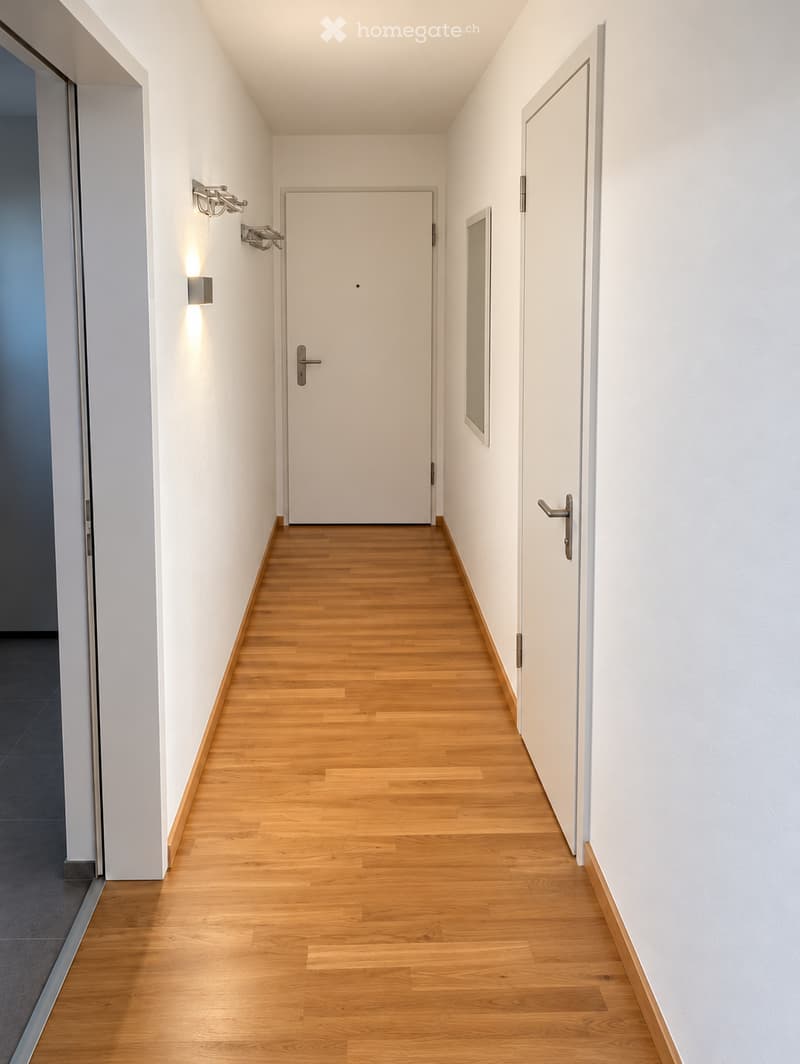 Clean, modern hallway with light wooden flooring, white walls, and several doors. Features wall-mounted light fixtures and a sleek design.