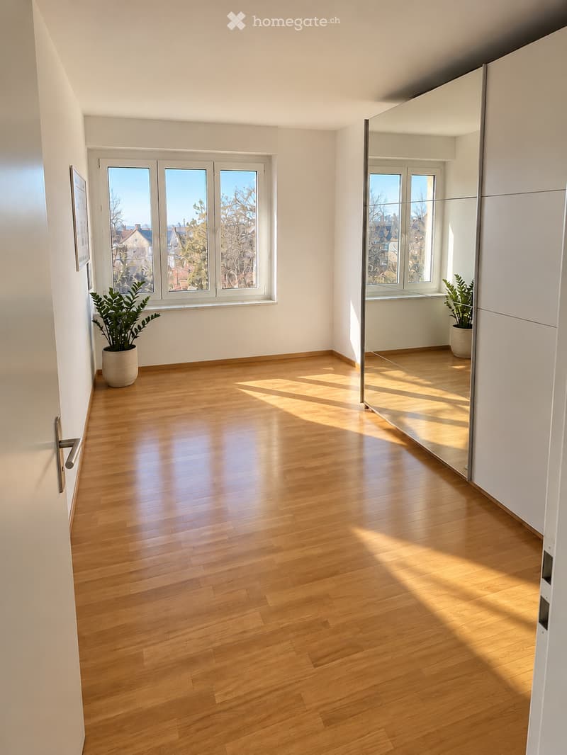 Empty room with wooden flooring, large windows, and white walls. Sunlight streams in, casting shadows on the floor. There are two potted plants near the windows and a mirrored closet on the right side.