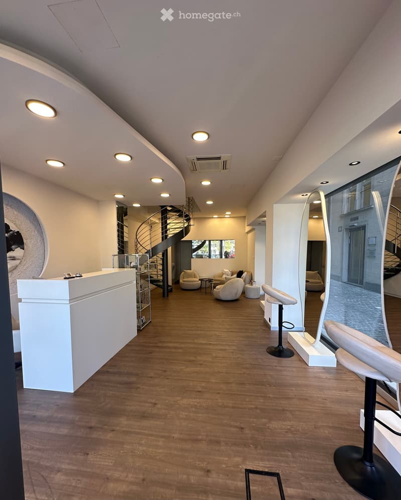 Modern reception area with wooden flooring, white reception desk, seating area with sofas and chairs, tall mirror, and a spiral staircase leading to an upper level.
