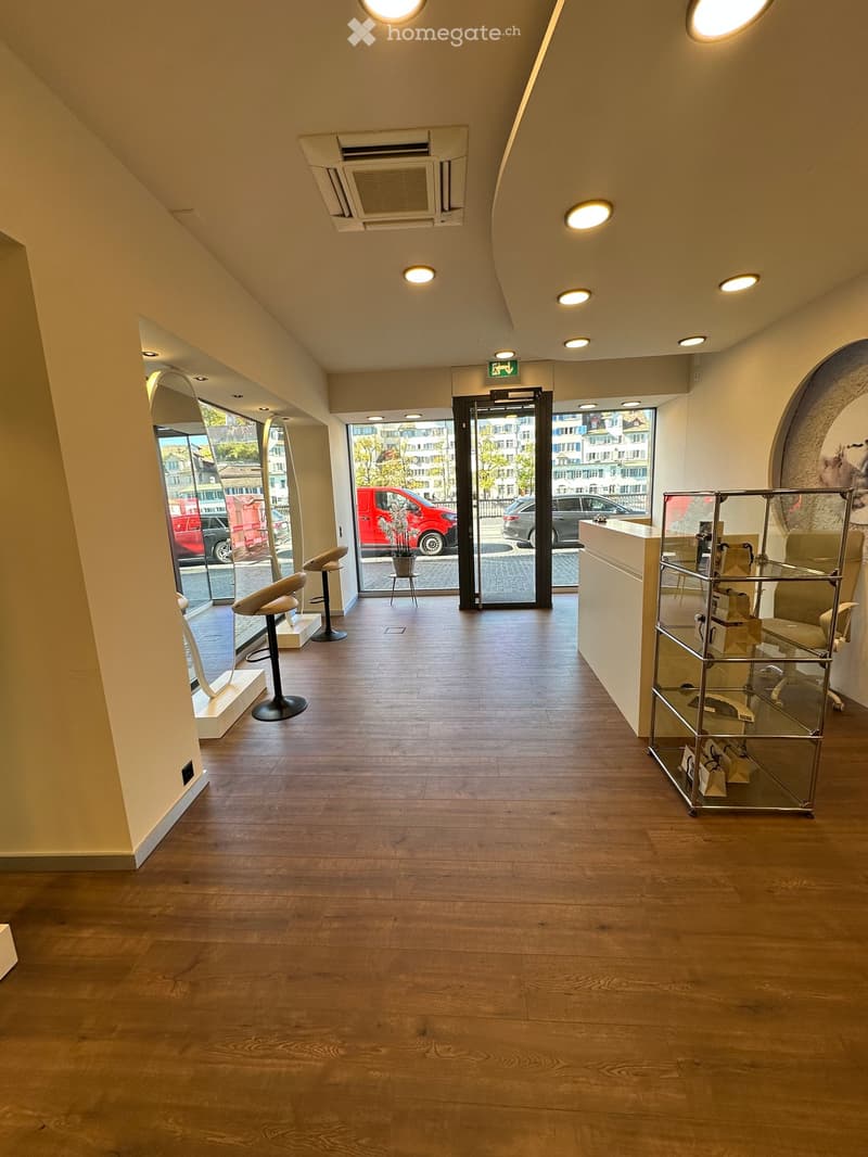 Modern reception area with wooden flooring, white walls, and recessed lighting. Features a white counter, glass display stand, and two white stools near the entrance.