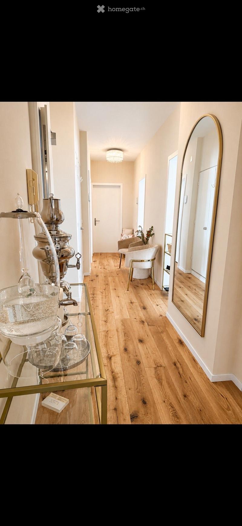 Elegant hallway with wooden flooring, a large oval mirror, a small seating area with a white armchair and side table, and a glass coffee cart with a coffee machine and glassware.