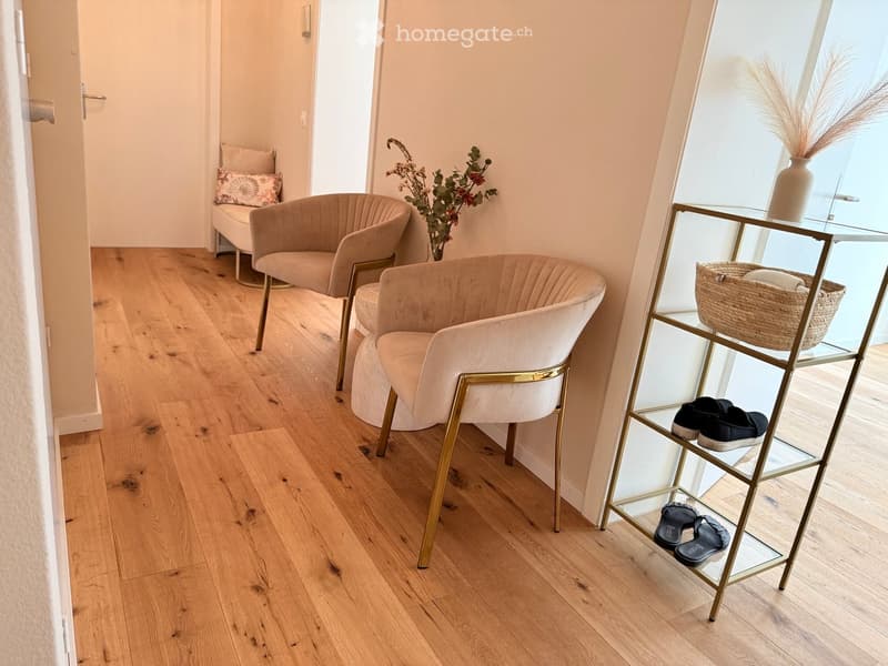 A small, well-lit room with light wooden flooring, three upholstered chairs, a glass shoe rack, and decorative vases.