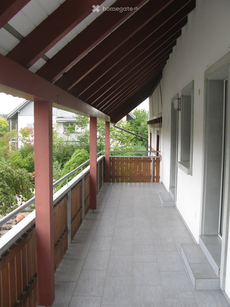 Balcony with tiled floor, wooden railing, and covered roof supported by red pillars. The view includes greenery and neighboring buildings.