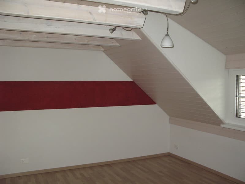 Empty attic room with wooden flooring, white walls, exposed wooden beams, and a single hanging light fixture.