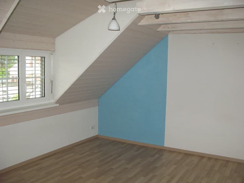 Empty attic room with wooden flooring, white walls, and a blue accent wall. Features a single window with white frame and a hanging light fixture.