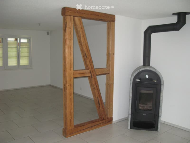 Empty room with tiled floor, white walls, large window with shutters, and a wood-burning stove with a chimney.