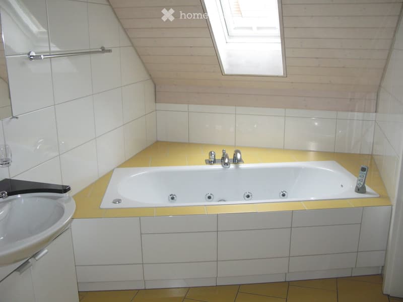 Modern bathroom with a large bathtub, sink, mirror, and a remote control placed on the edge of the tub. The walls are tiled in white and light beige, and the floor is yellow.