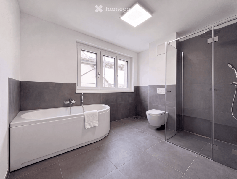 Modern bathroom with a bathtub, toilet, glass-enclosed shower, and a window providing natural light.